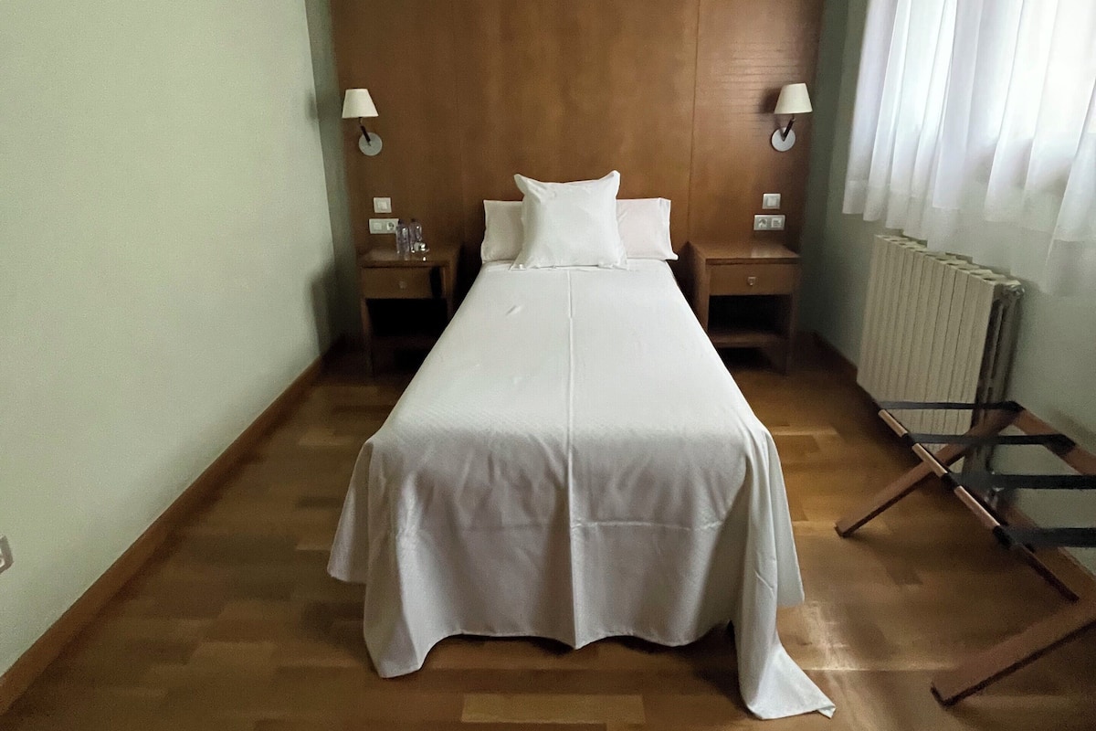 Quarto de hotel com uma cama de solteiro, cabeceira de madeira, duas mesas de cabeceira com luminárias, um radiador e uma janela com cortinas.