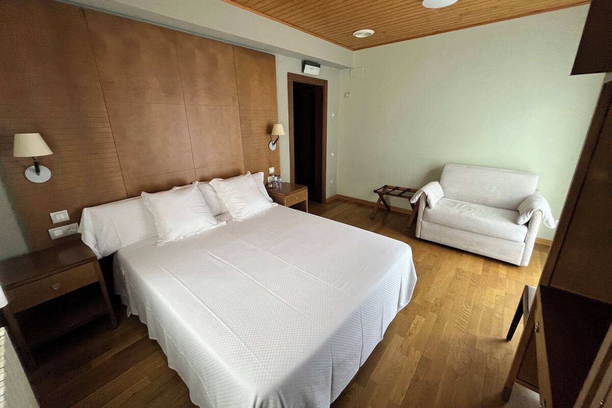 Quarto de hotel com uma cama grande, uma poltrona branca, teto de madeira e uma mesa pequena.