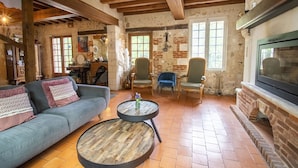 A rustic living room with exposed brick walls, a fireplace, and a large TV. There is a sofa, two chairs, and a round coffee table. - Le Prieure des Fontaines (Les Preaux)
