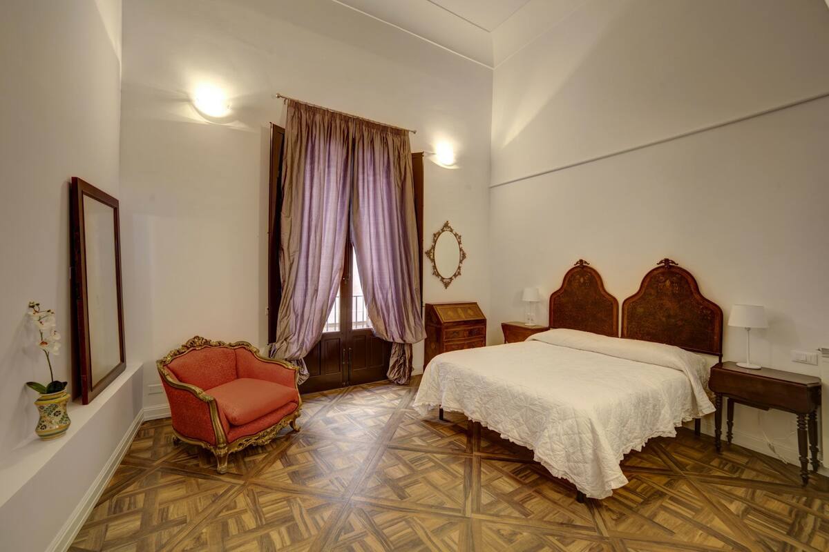 A bedroom with a wooden floor, a bed with a white bedspread, a red armchair, a wooden nightstand, a mirror, and a window with curtains.