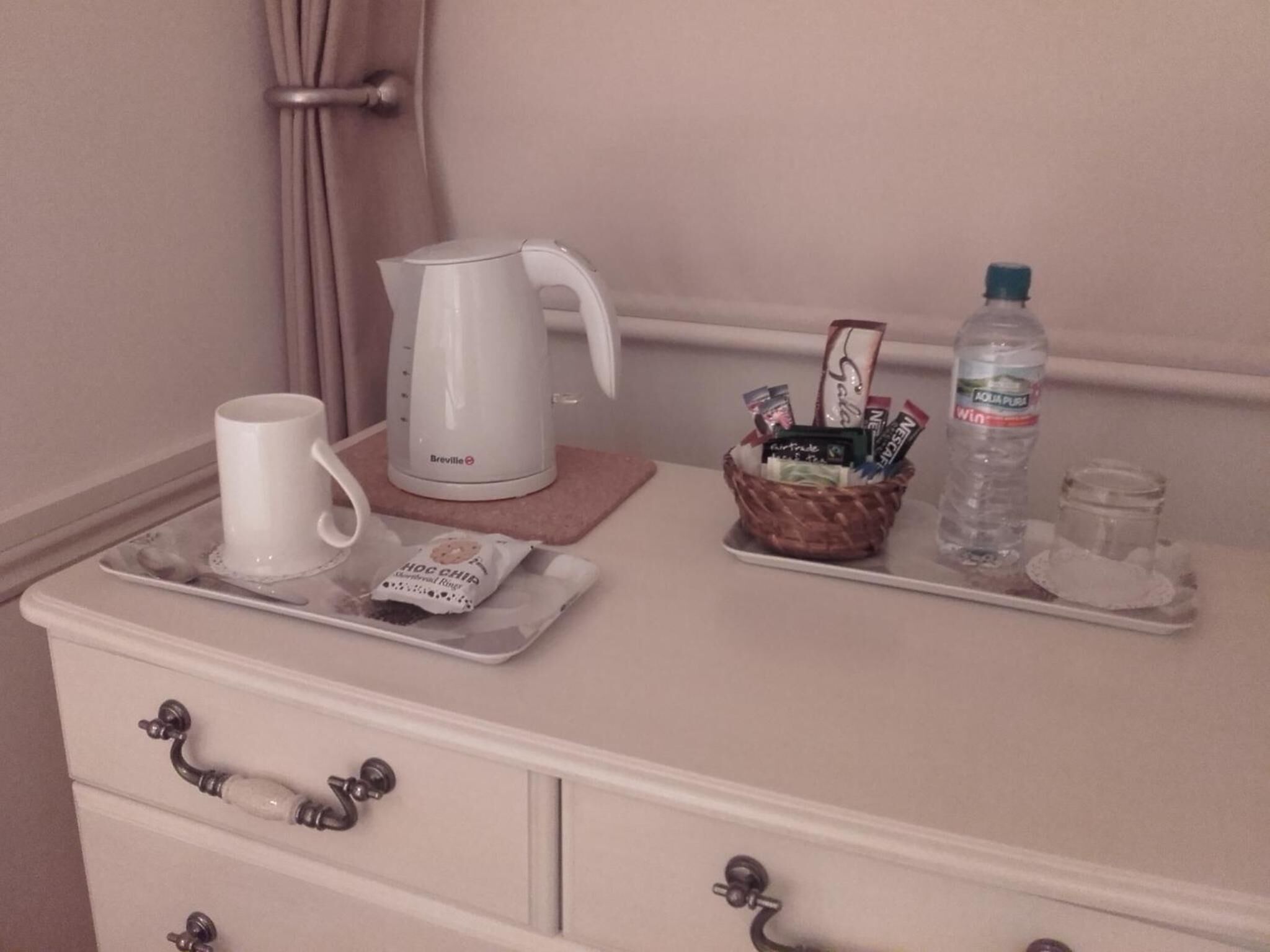 Een dienblad met een witte mok, een Breville koffiezetapparaat, een mand met snacks, een waterfles en een glas water op een witte dressoir.