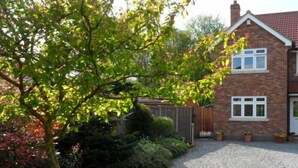 Une maison à deux étages, construite en briques, avec un toit recouvert de tuiles rouges, une porte d’entrée et un garage. On y trouve des plantes en pot et une allée gravillonnée.