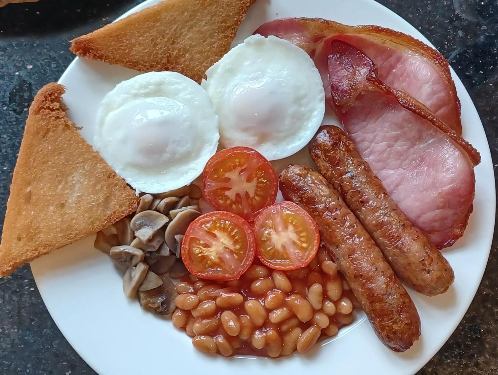 Un plato de comida para el desayuno que incluye huevos, tocino, salchichas, tostadas, frijoles y tomates.