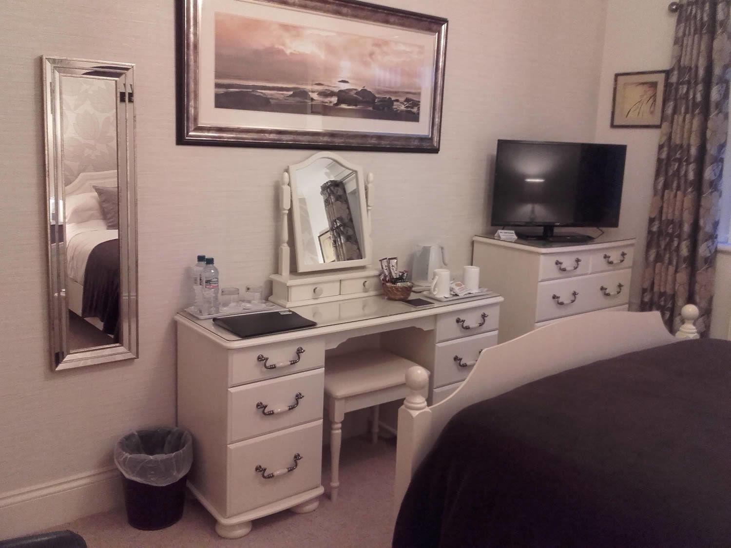 A hotel room with a bed, a desk with drawers, a mirror, a television, and a framed picture on the wall.