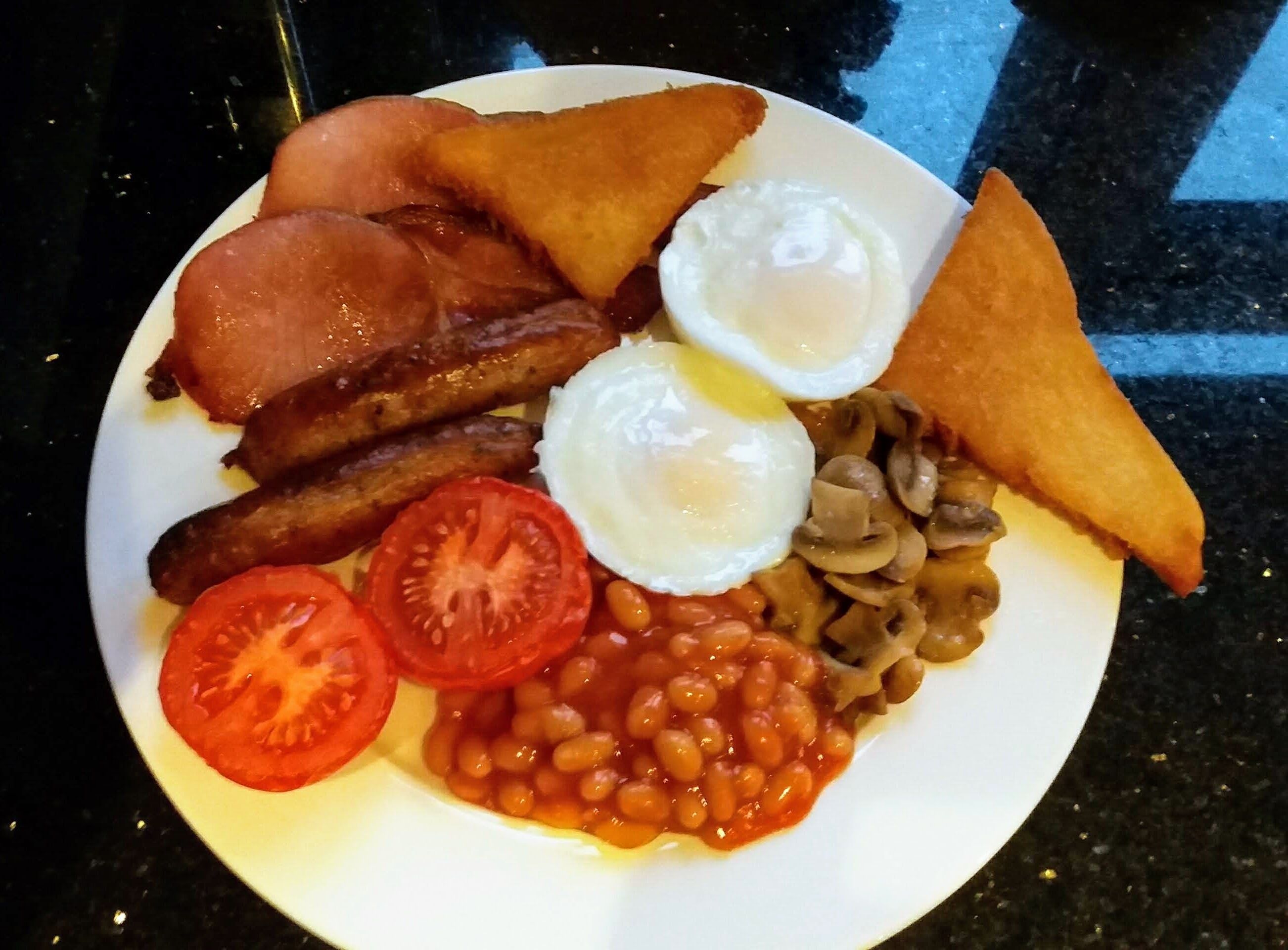 A plate of breakfast food including eggs, bacon, sausages, mushrooms, beans, and tomatoes.