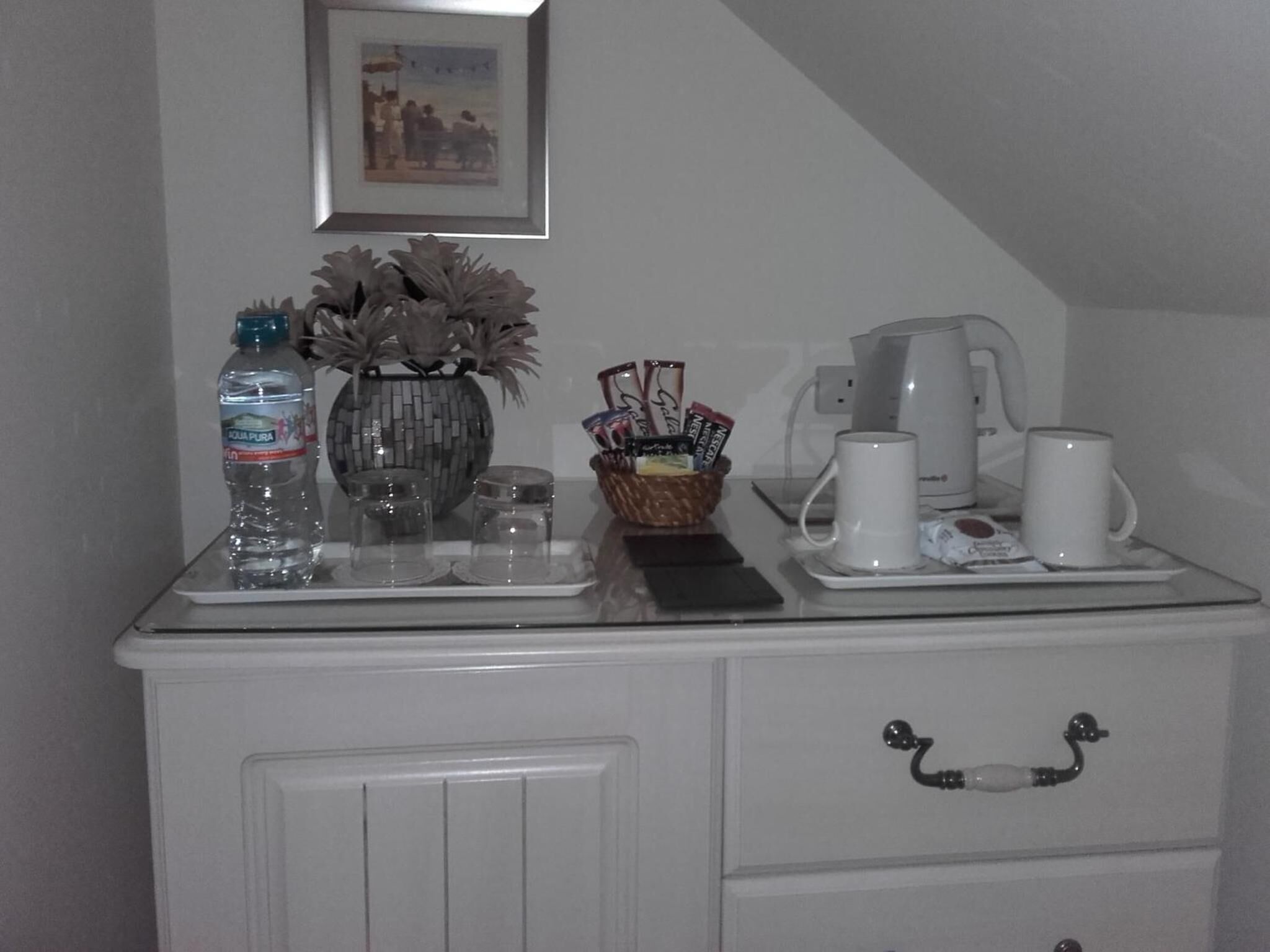 A kitchen counter with a coffee maker, two mugs, a basket of snacks, and a water bottle.