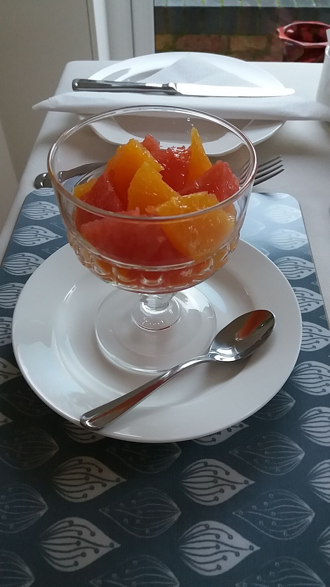 Un dessert aux fruits servi dans un bol en verre, posé sur une assiette blanche avec une cuillère à côté, le tout placé sur une table recouverte d’une nappe à motifs bleus.