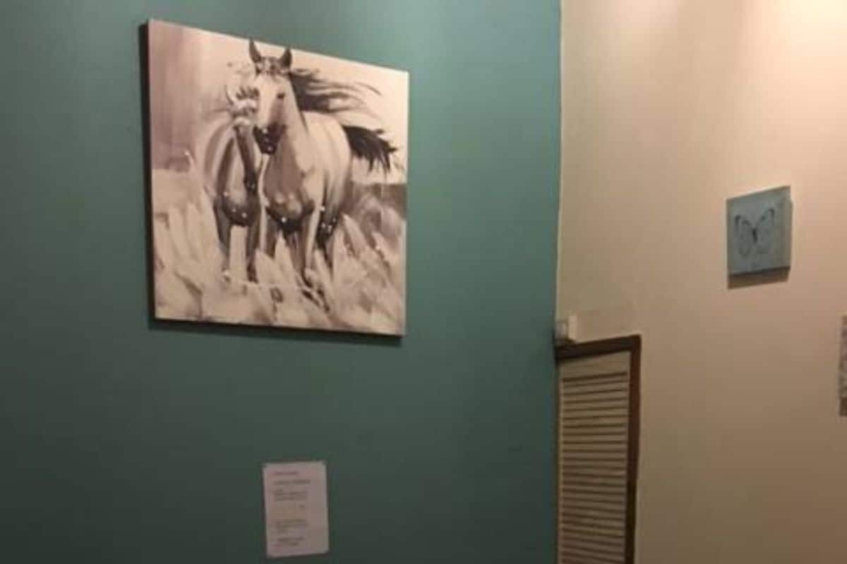 A room with a bed, a small table with a cup, a painting of horses, and a radiator.