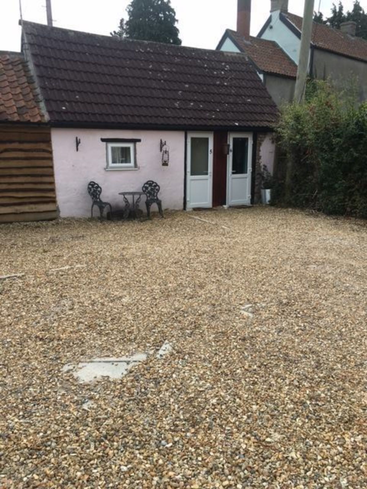 A small building with a tiled roof, a door, and a window. There is a gravel driveway and two chairs outside.