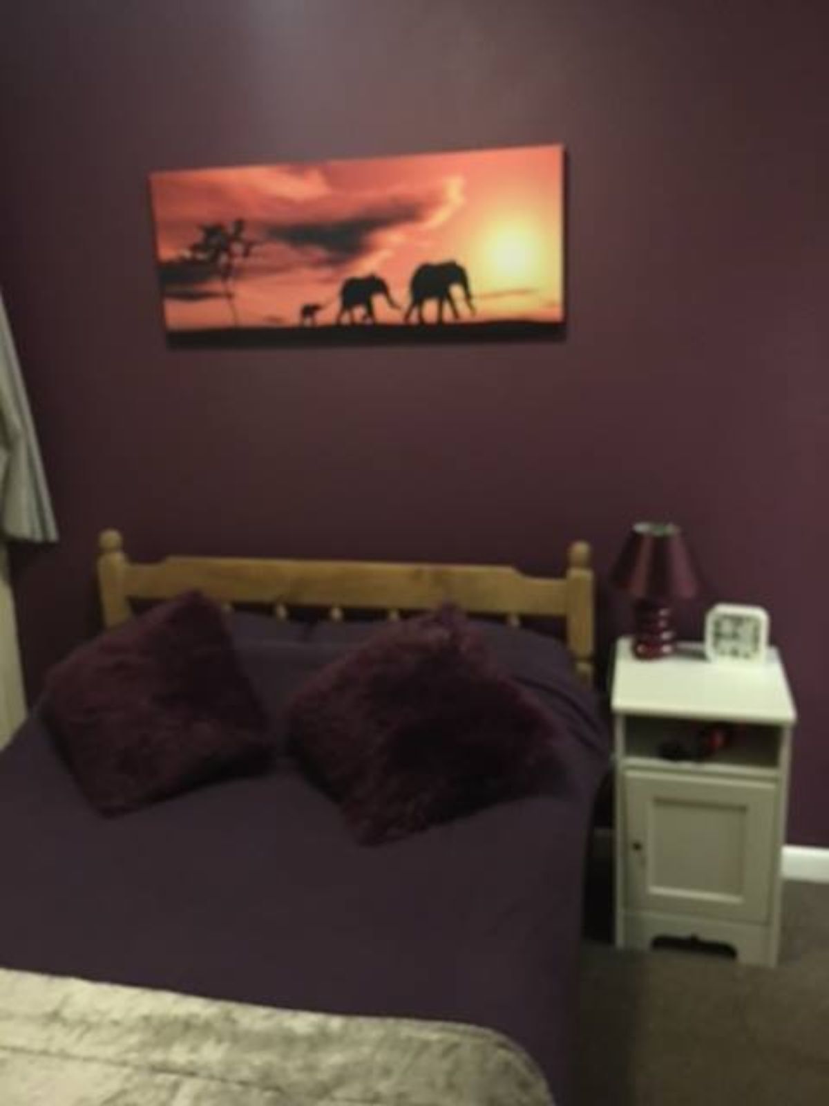 A bedroom with a bed, pillows, a nightstand with a lamp, and a painting on the wall.