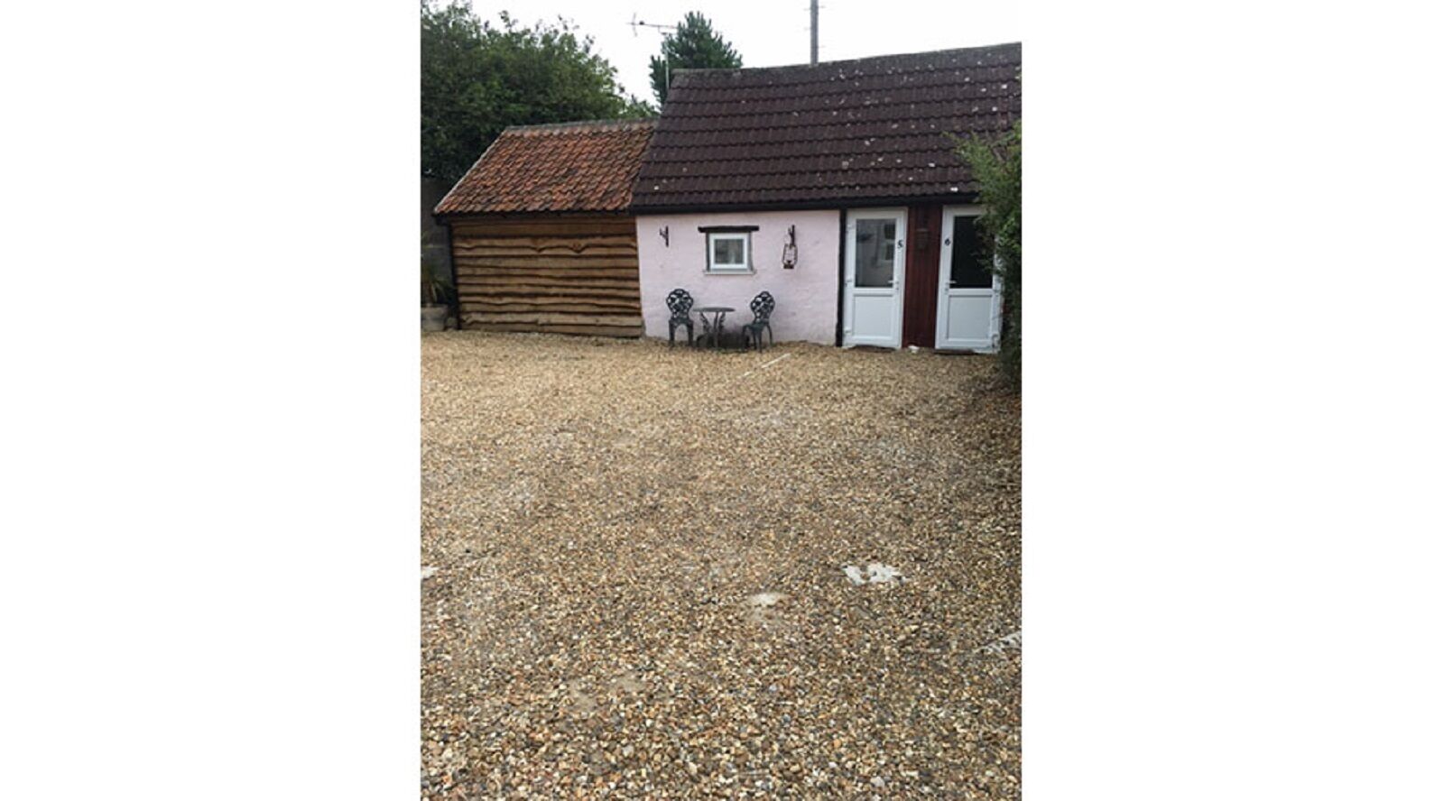 A rustic building with a tiled roof, a wooden structure to the left, and a small patio area with two chairs and a table.