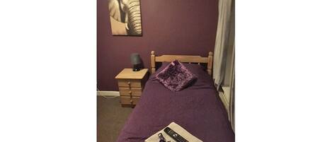 A single bed with a purple blanket, a wooden nightstand with a lamp, and a wall-mounted picture.