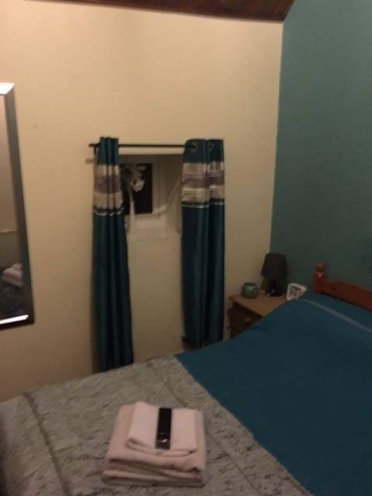 A bedroom with a bed, a window with curtains, a bedside table with a lamp, and a wall-mounted shelf.