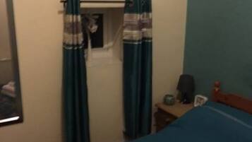 A bedroom with a bed, a window with curtains, a bedside table with a lamp, and a wall-mounted shelf.