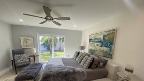 Un dormitorio con una cama, una silla, un banco, un ventilador de techo, un cuadro y vistas al exterior.