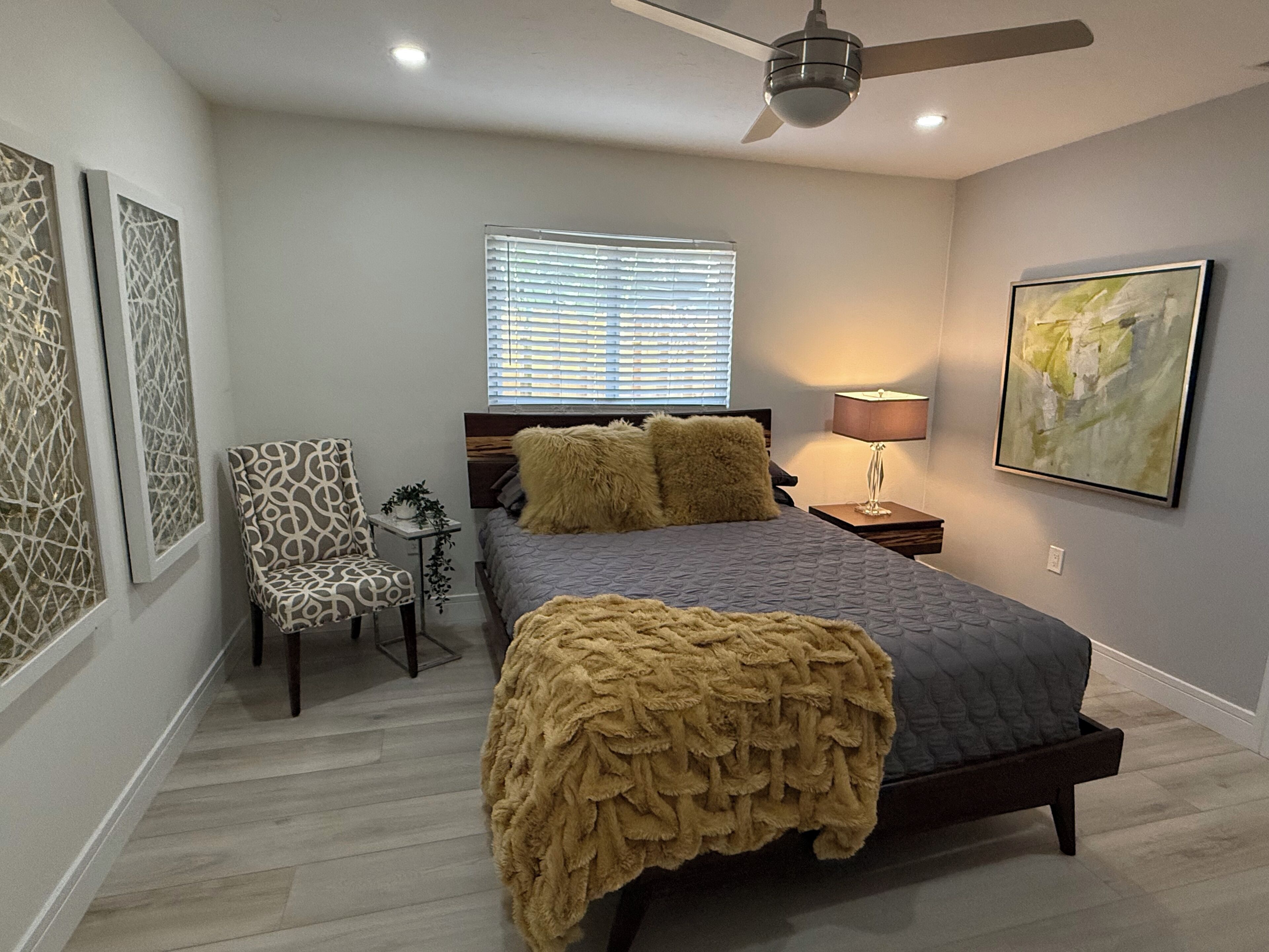 A bedroom with a bed, a chair, a window with blinds, a ceiling fan, a lamp, and artwork on the walls.
