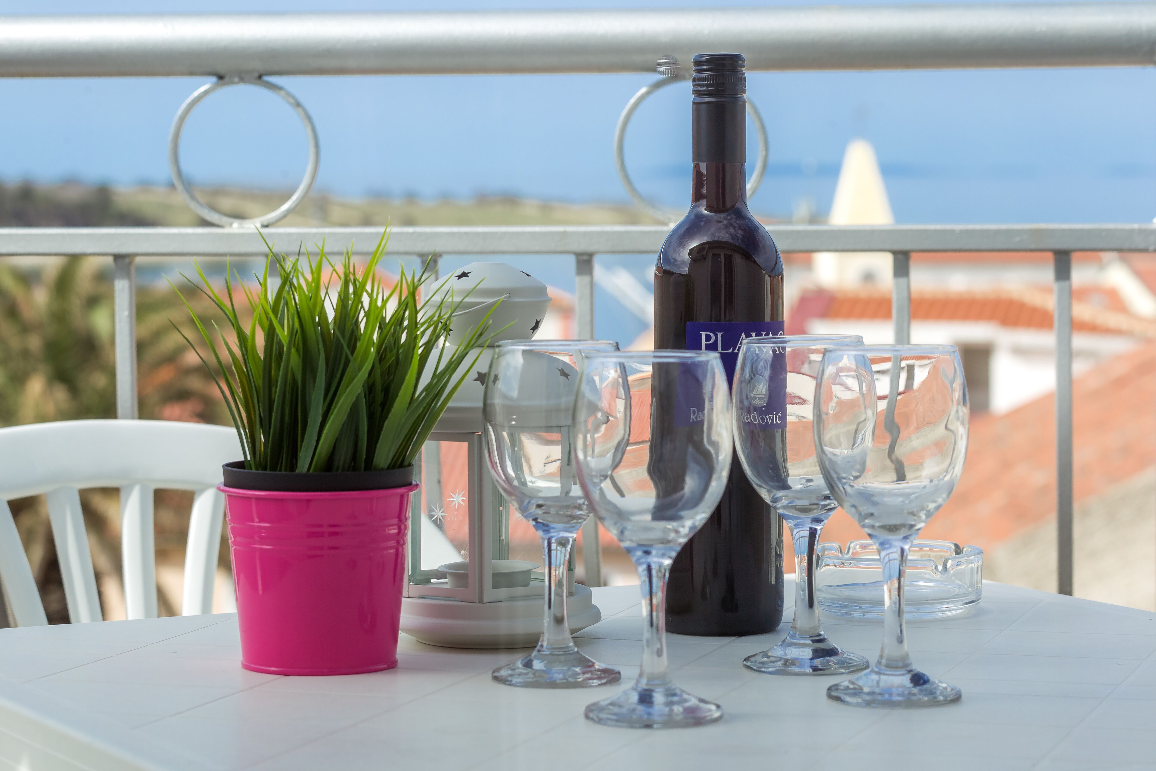 A balcony with a table set for dining, featuring a bottle of wine, wine glasses, and a potted plant.