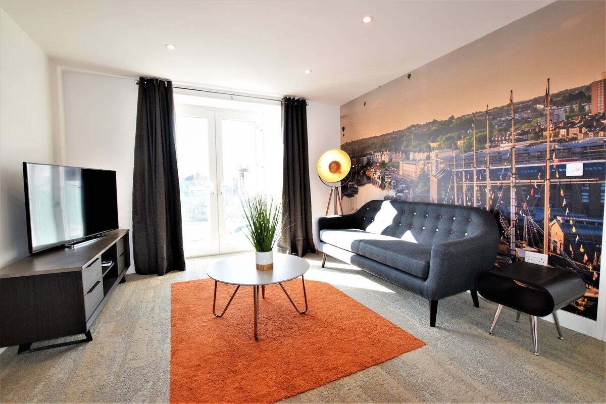 A modern living room with a flat-screen TV, a grey sofa, a coffee table with a plant, and a cityscape mural on the wall.