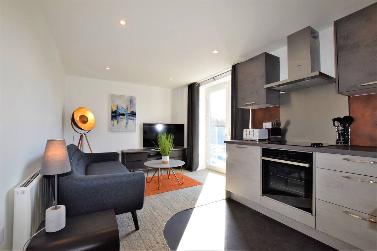 A modern living room with a sofa, a coffee table, and a TV. There is a kitchen with a stove, oven, and cabinets.