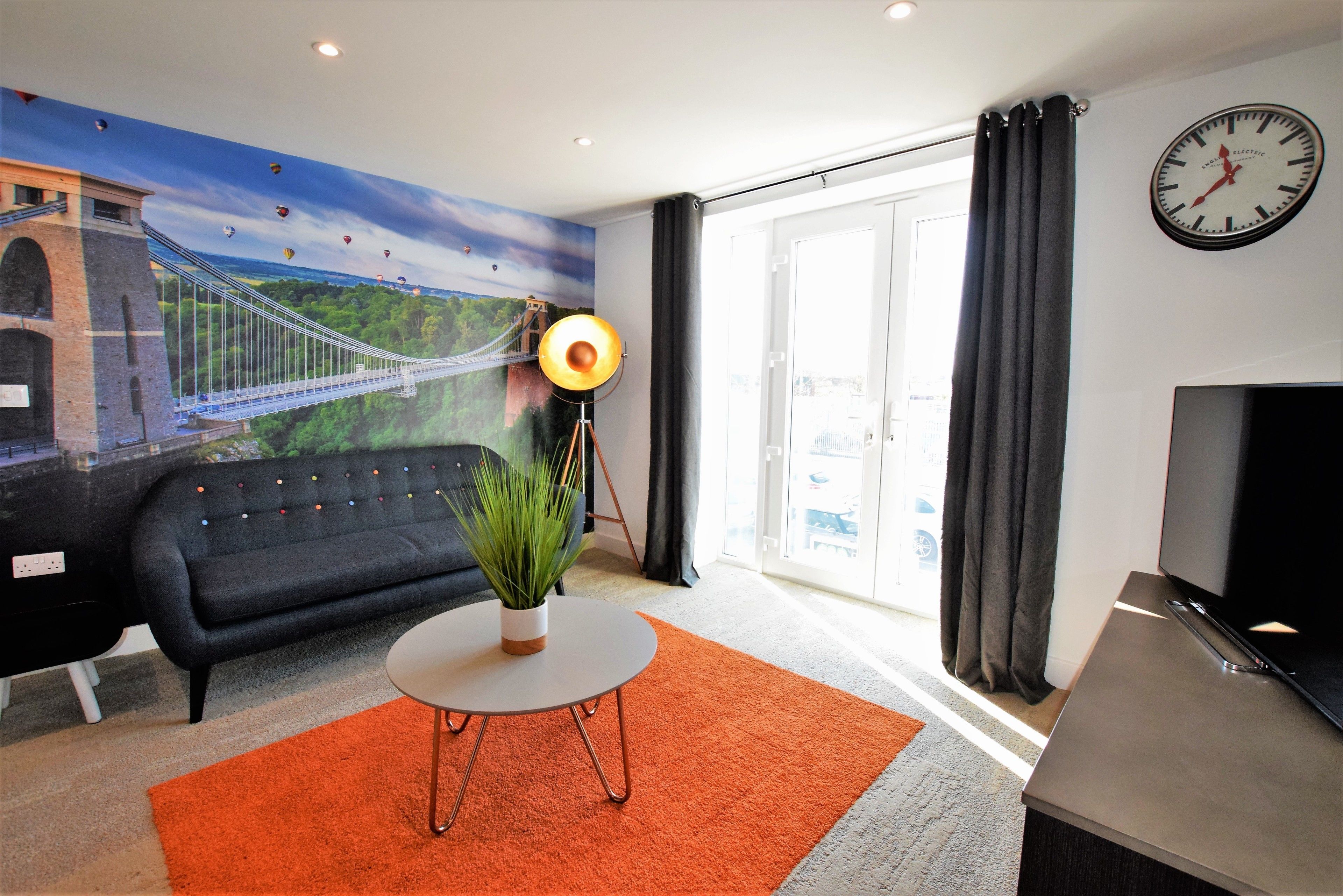 A living room with a sofa, a round table with a plant, a TV, a clock, and a mural of a bridge.