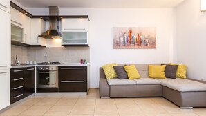 A modern kitchen with dark cabinets, a built-in oven, and a stainless steel range hood. A gray sectional sofa with yellow and gray cushions is positioned against the wall. - Apartment with Terrace and Partial Sea View and grill (Novalja)