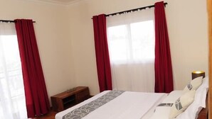 Un dormitorio con cama, piso de madera, cortinas, una lámpara de noche y una mesita de madera.