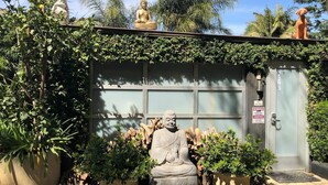 Um edifício com uma grande estátua de Buda em frente, rodeado por plantas em vasos e uma porta de garagem.