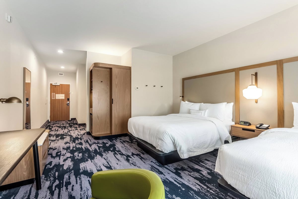 A hotel room with a bed, bedside table, and a green chair. There is a wooden wardrobe and a door in the background.