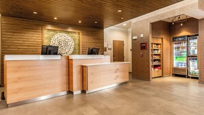 A reception area with wooden reception desks, a decorative wall piece, a refrigerator stocked with beverages, and a fire extinguisher on the wall.