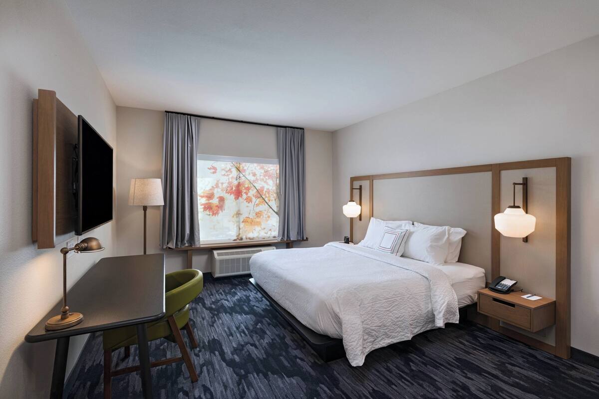 A hotel room with a bed, bedside table, lamp, desk, chair, and a window with curtains.