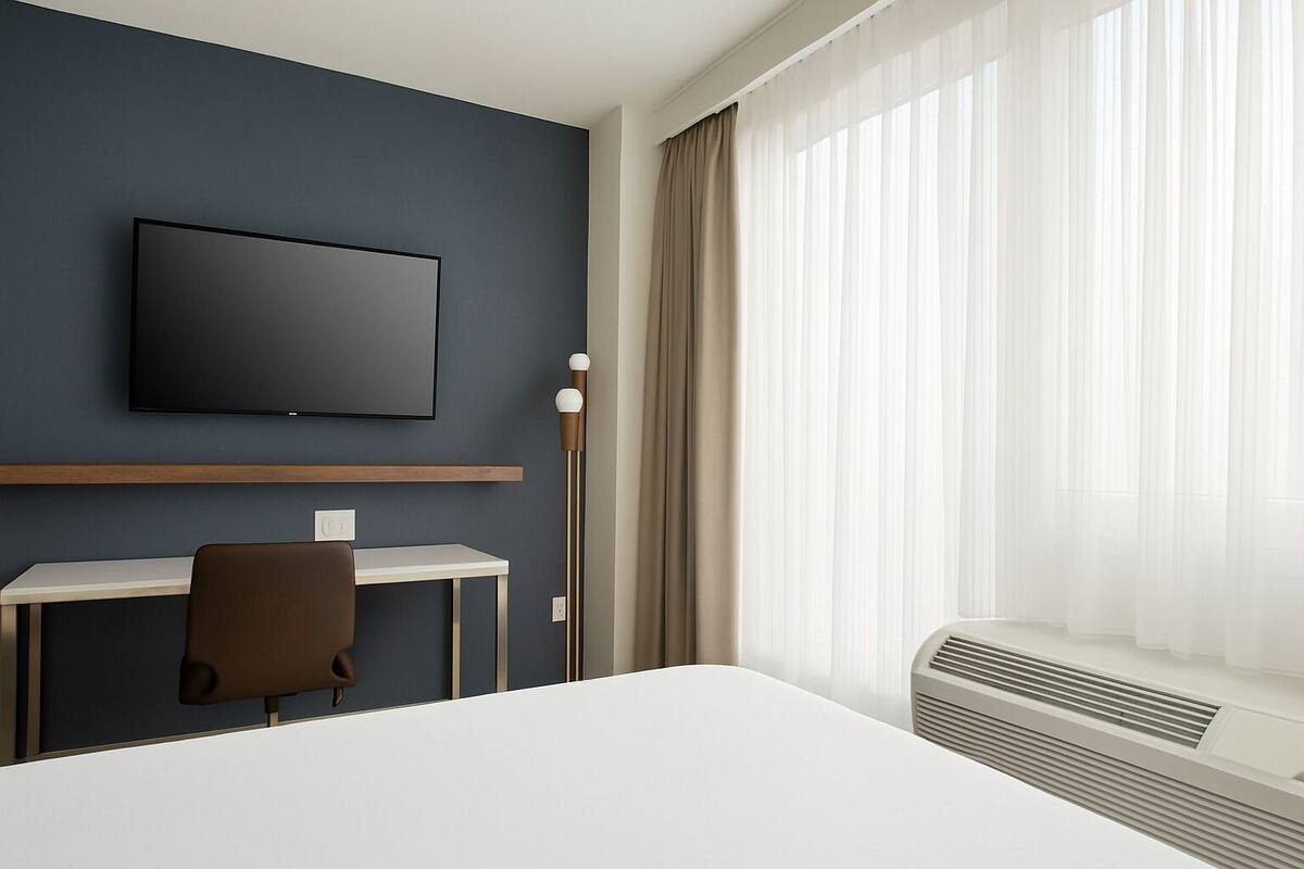 Une chambre d’hôtel dotée d’un téléviseur à écran plat fixé au mur, d’un bureau avec une chaise, d’une grande fenêtre avec des rideaux légers et d’un système de climatisation intégré.