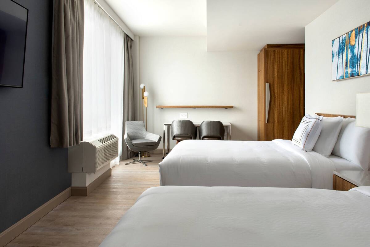 Une chambre d’hôtel avec deux lits, un bureau avec des chaises, une grande fenêtre avec des rideaux, un téléviseur fixé au mur et une armoire en bois.