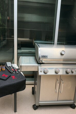 Una parrilla portátil Weber con una mesa colocada adelante, que incluye una botella de sazonador, una espátula y un tenedor.