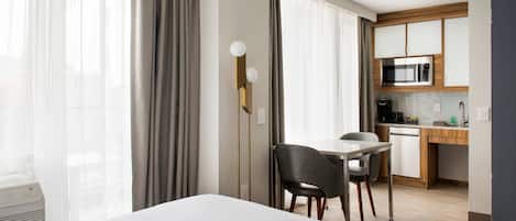 A hotel room with a bed, a desk, a chair, a kitchenette, and large windows with curtains.