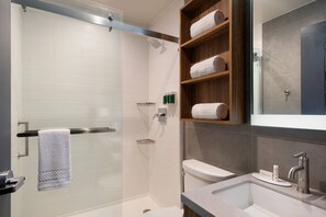 Un baño moderno con ducha de vidrio, un estante de madera con toallas, un lavamanos con espejo y un inodoro.