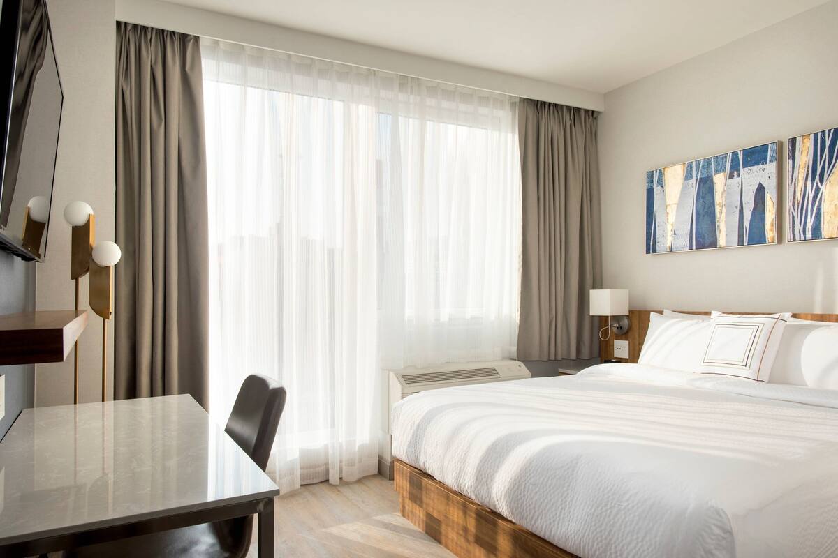 Une chambre d’hôtel avec un lit, un bureau, une chaise, une télévision et une grande fenêtre avec des rideaux.
