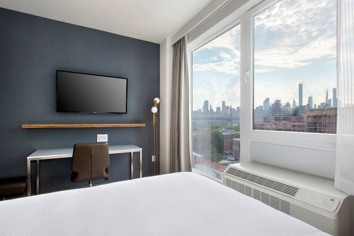 Une chambre d’hôtel équipée d’un lit, d’un bureau, d’une chaise, d’un téléviseur fixé au mur et d’une grande fenêtre donnant sur la ville.