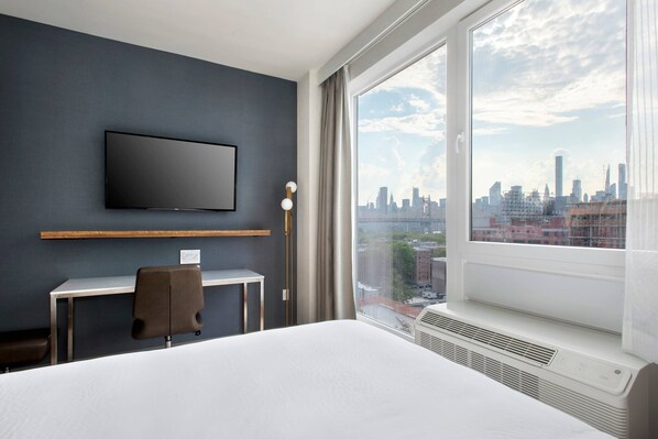 A hotel room with a bed, a desk, a chair, a TV mounted on the wall, and a large window offering a city view.