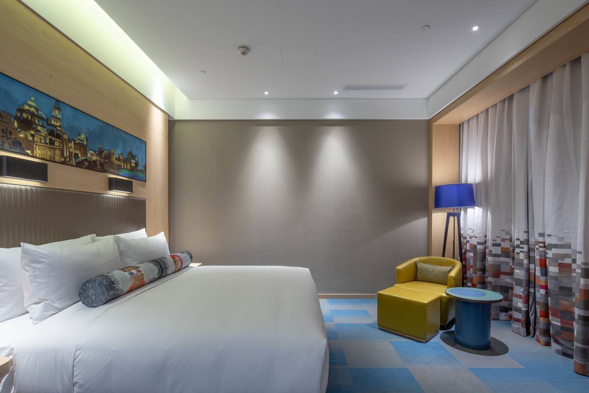 A hotel room with a large bed, a yellow armchair, a small round table, and a painting of a cityscape on the wall.