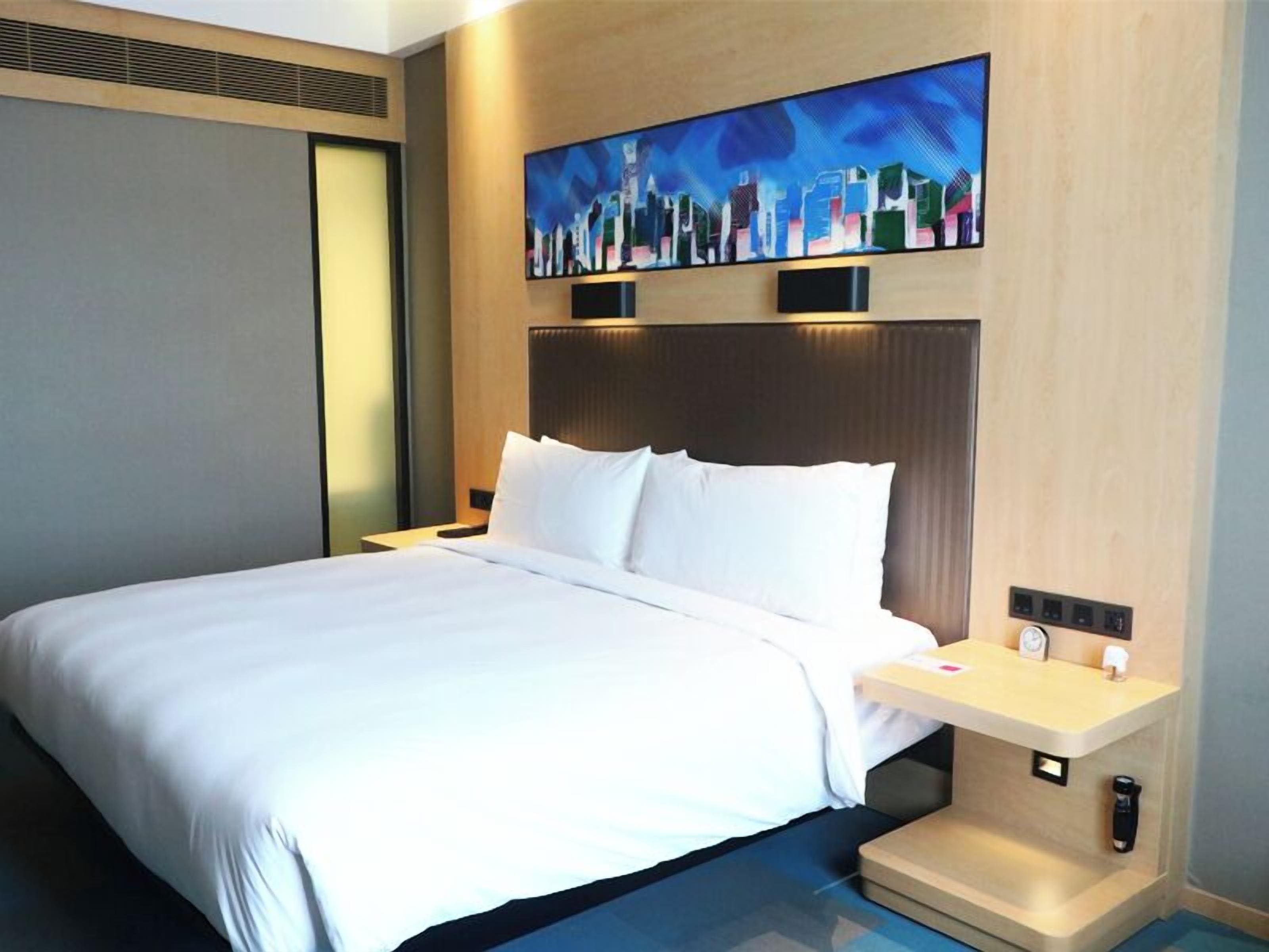 A hotel room with a large bed, a nightstand, a painting on the wall, and a cityscape view.