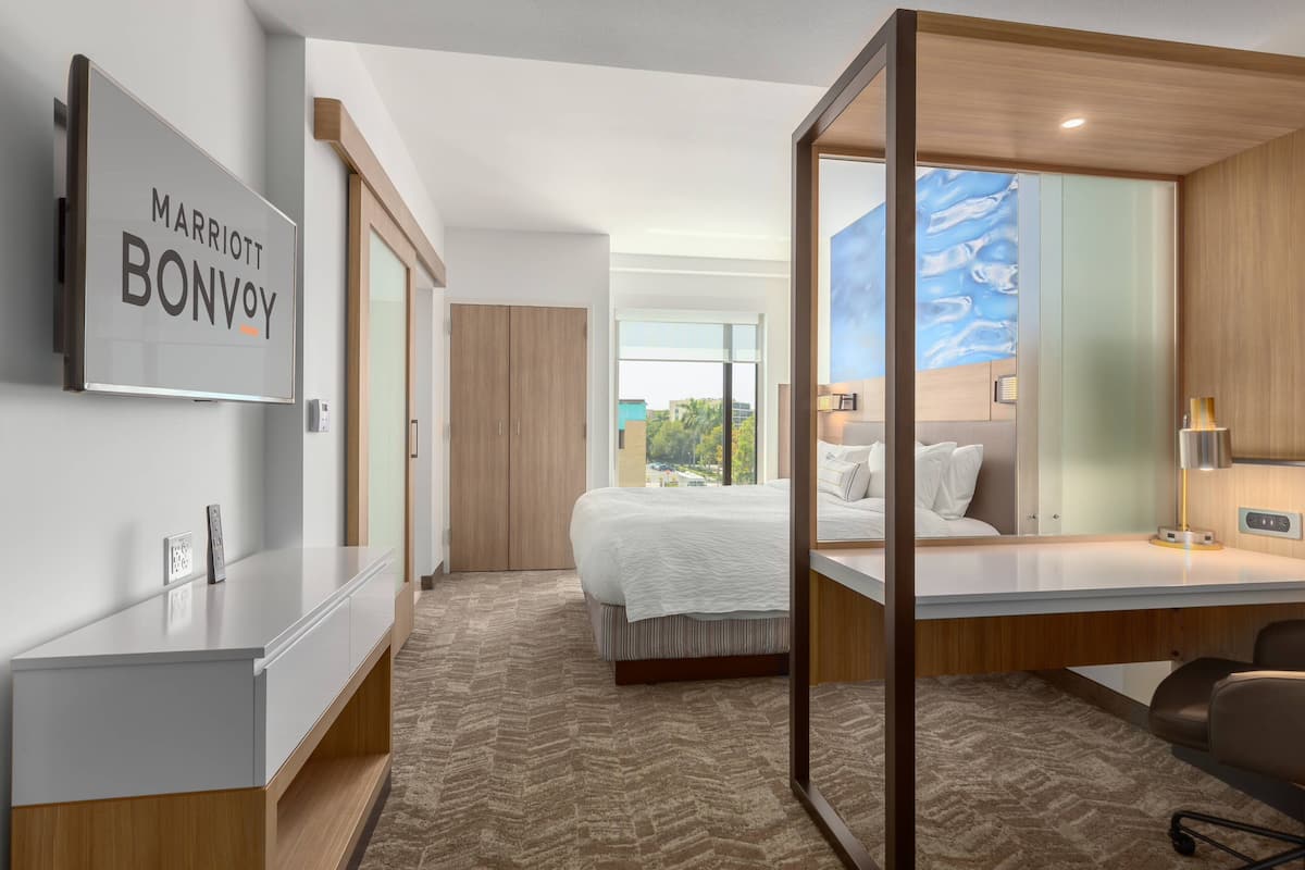 Habitación de hotel moderna con una cama grande, un escritorio con computadora, un espejo amplio y vistas al exterior.