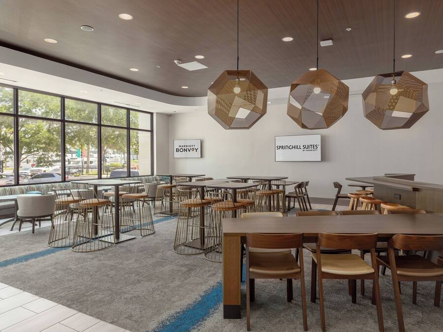 A modern hotel lobby with large windows, hexagonal hanging lights, and a variety of seating arrangements.