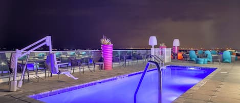 Zona de piscina en la azotea con piscina iluminada en azul, sillas de descanso y vista del horizonte urbano de noche.