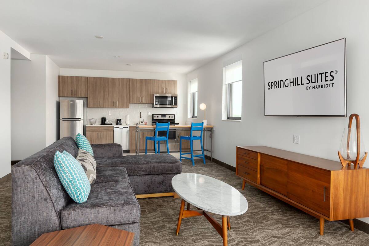 Una sala de estar moderna con un sofá modular gris, una mesita de centro de madera y un comedor con sillas azules. Se ve una cocina con armarios de madera y electrodomésticos de acero inoxidable al fondo.