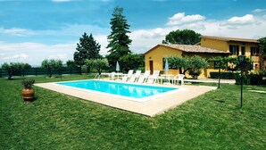 Una piscina con lettini, circondata da un prato curato e un edificio con tetto in tegole.