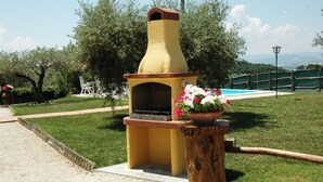 Forno in mattoni all'aperto con un vaso di fiori su un ceppo di legno, circondato da un'area erbosa con piscina e recinzione in legno.