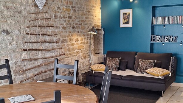 A dining area with a round wooden table and chairs, a stone wall, a blue wall with a bookshelf, and a sofa with patterned cushions. - La Maison de la Grève (Ouistreham)