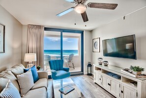 Un soggiorno con una grande TV, una ventola a soffitto, un divano, una poltrona e vista sull'oceano.