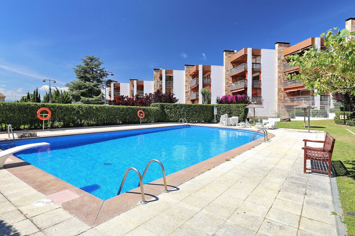 A swimming pool with a lifebuoy and no swimming sign, surrounded by a paved area with a bench and lounge chairs.