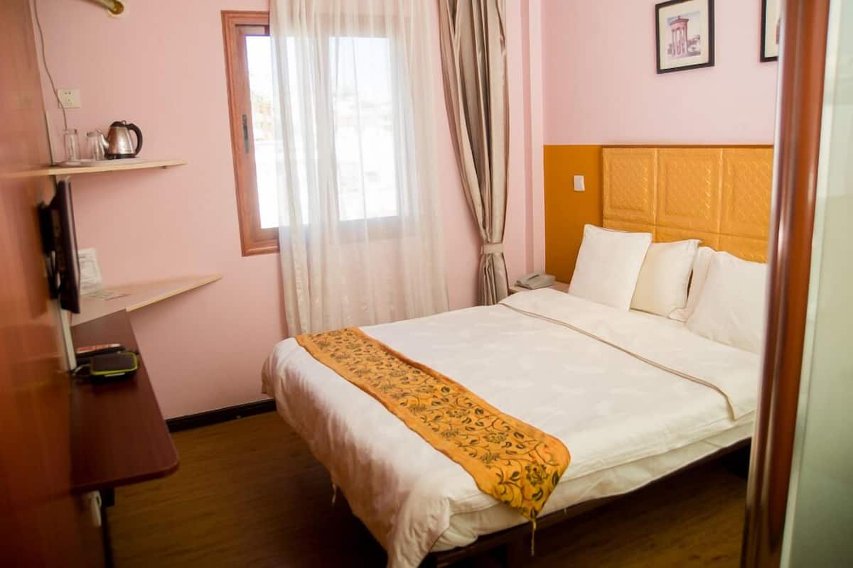 A hotel room with a bed, a wooden nightstand, a window with sheer curtains, and a wall-mounted shelf with a kettle and glasses.